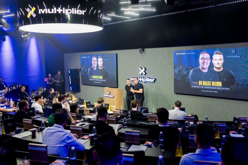 Plateia de empresários em evento presencial da Multiplier Educação, com telões exibindo os palestrantes e a frase "Um Brasil melhor através de empresas melhores"